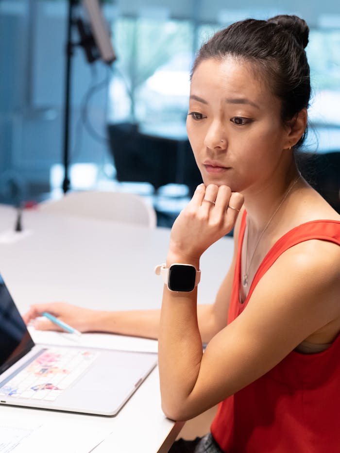 Asian woman in a red tank top working on a laptop indoors, wearing a smart watch.